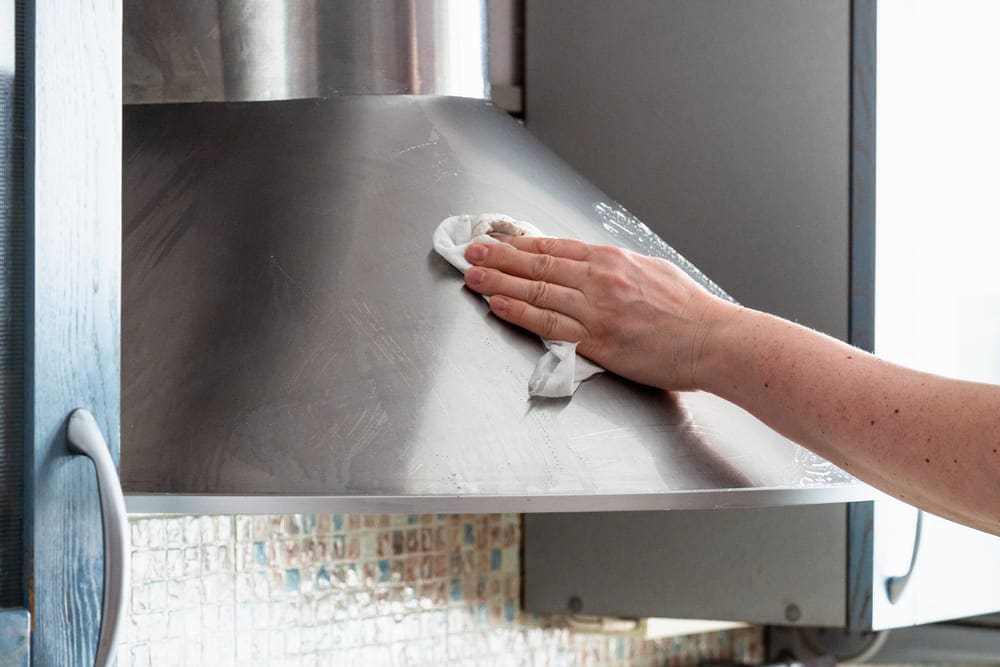 A hand with a rag, gently cleaning a range hood made of stainless steel.