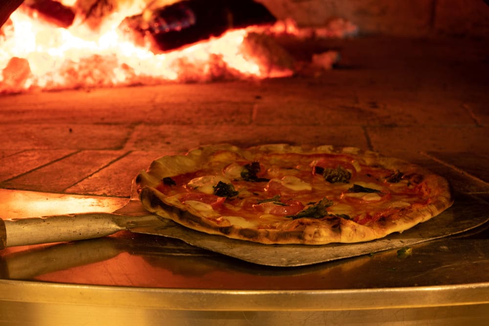 A person off camera, learning how to use a pizza oven by placing their first on a pizza peel onto the bricks