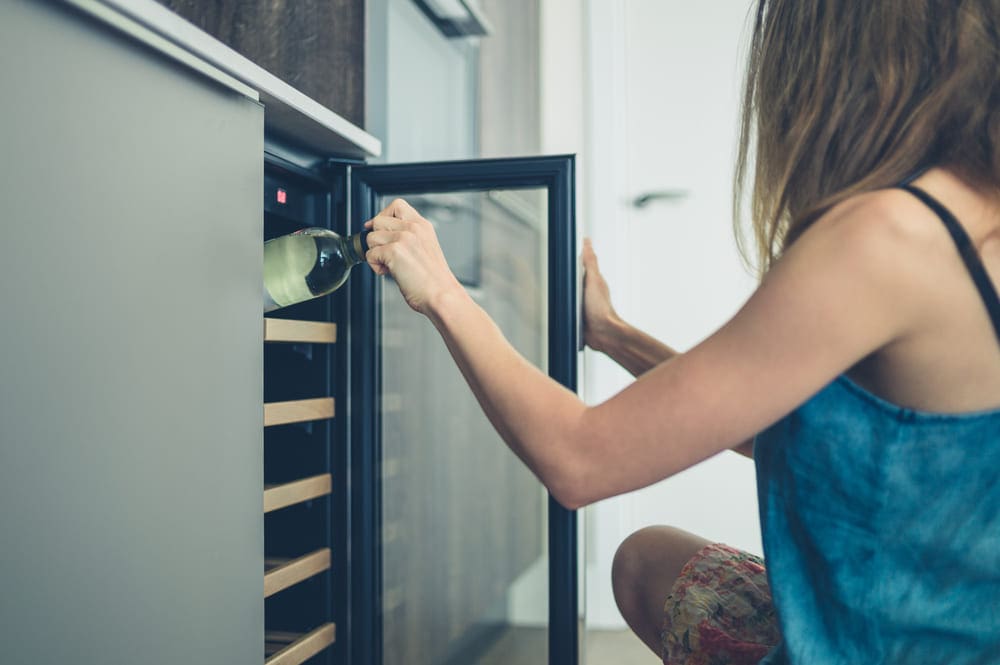 A young woman removes a bottle of wine from a wine cooler, an appliance known to keep consistency and best temperatures for storing wine.