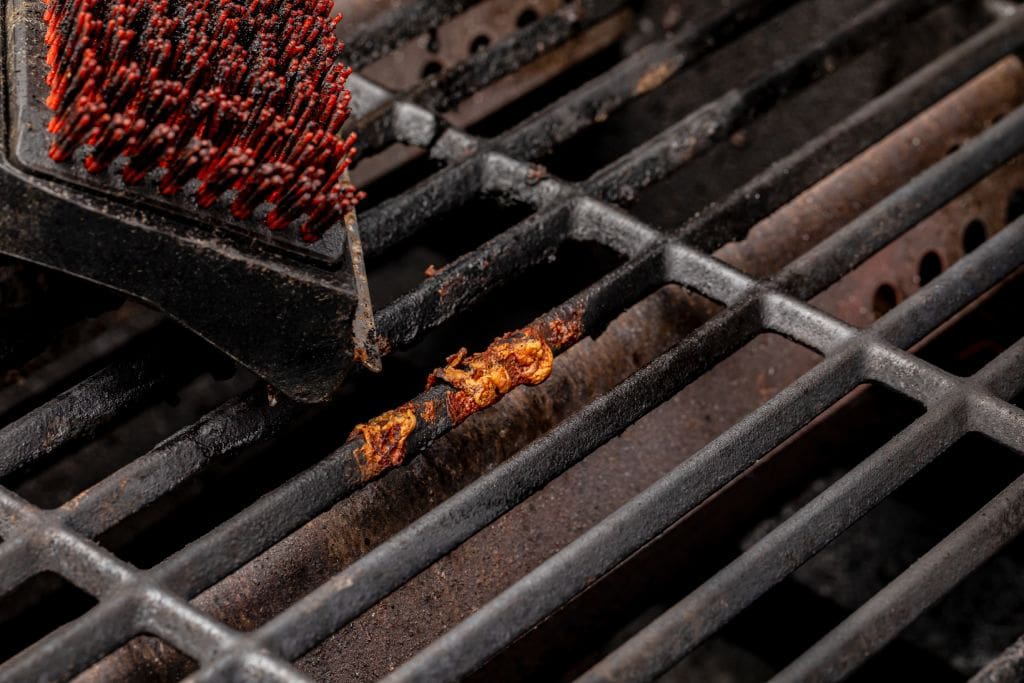 Close up of a grill brush scrubbing food residue from dirty grill grates to show how to clean a grill safely.