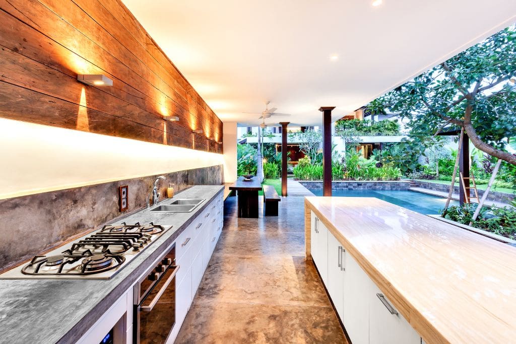 A modern outdoor kitchen, sleek concrete countertops, and wood accents beside a garden pool, illustrating outdoor kitchen cost ideas for a luxury home patio design.