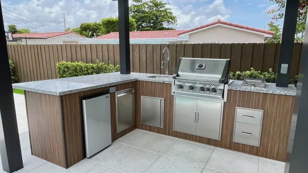 Modern outdoor kitchen with stainless steel grill, sink, and outdoor refrigerator drawers built into a stone countertop setup.