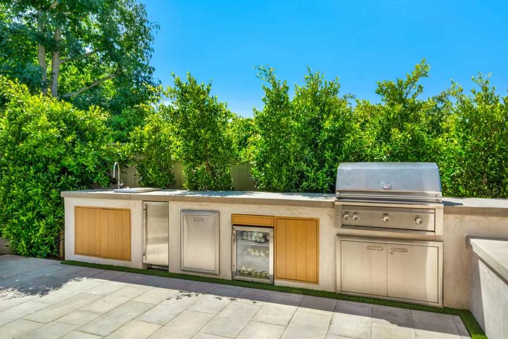 outdoor-kitchen-bbq Outdoor kitchen with built-in grill, storage, and outdoor refrigerator drawers surrounded by greenery under clear sky.